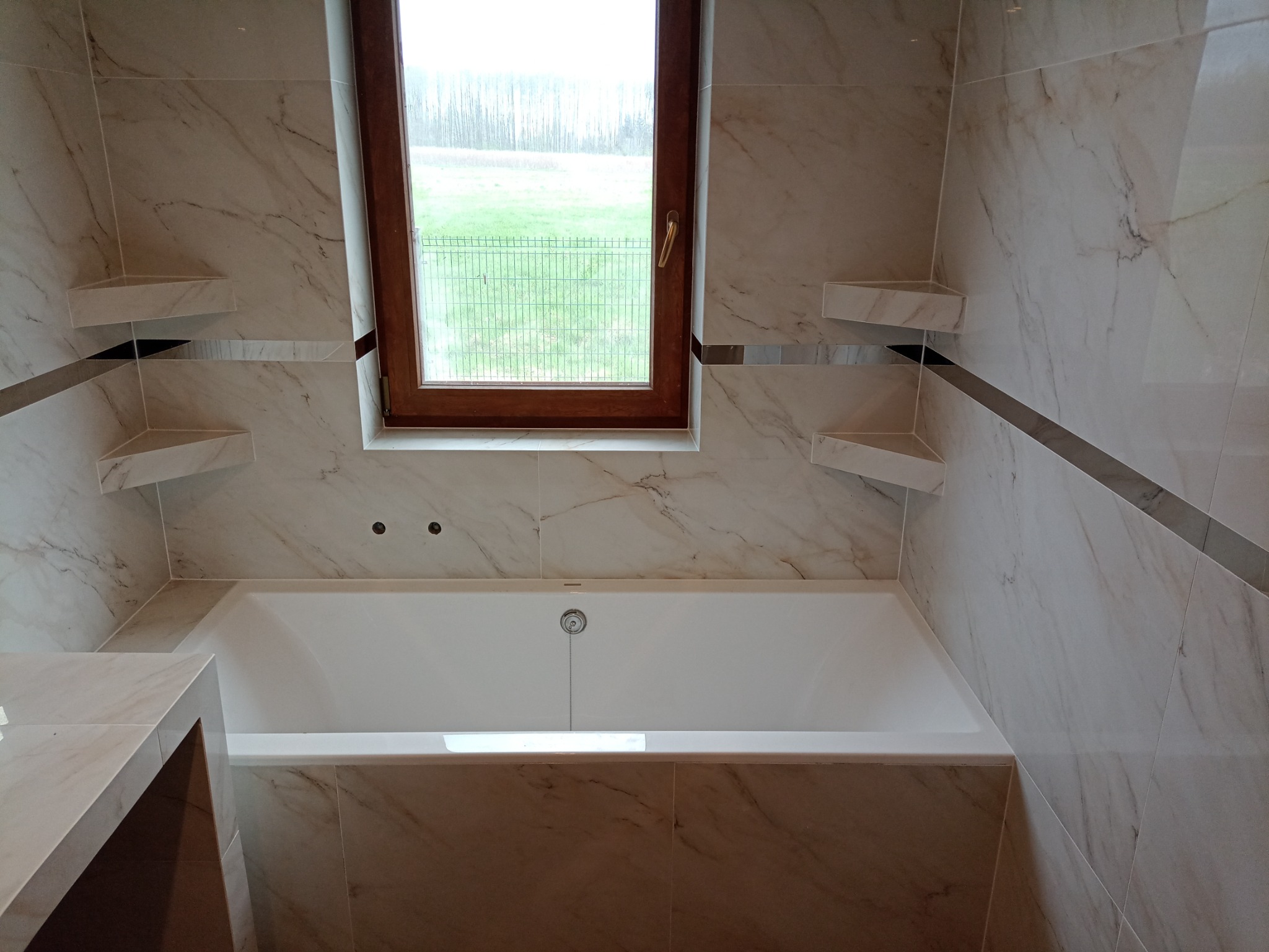 Wykończona łazienka z wanną, marmurowe płytki, drewniane okno z widokiem na zewnątrz i wbudowane półki narożne.