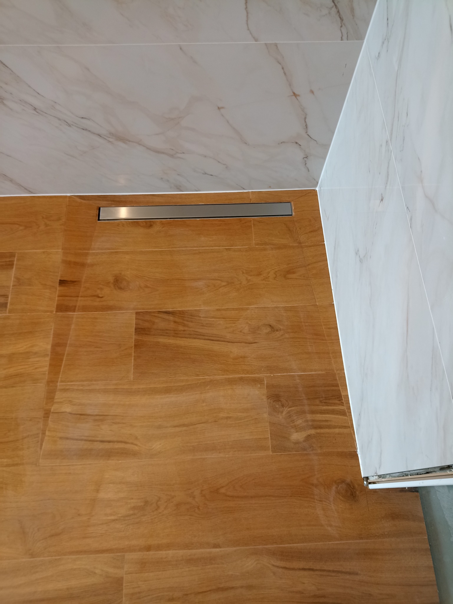 Wykończona łazienka z drewnopodobną podłogą i marmurowymi płytkami ściennymi oraz odpływem liniowym.