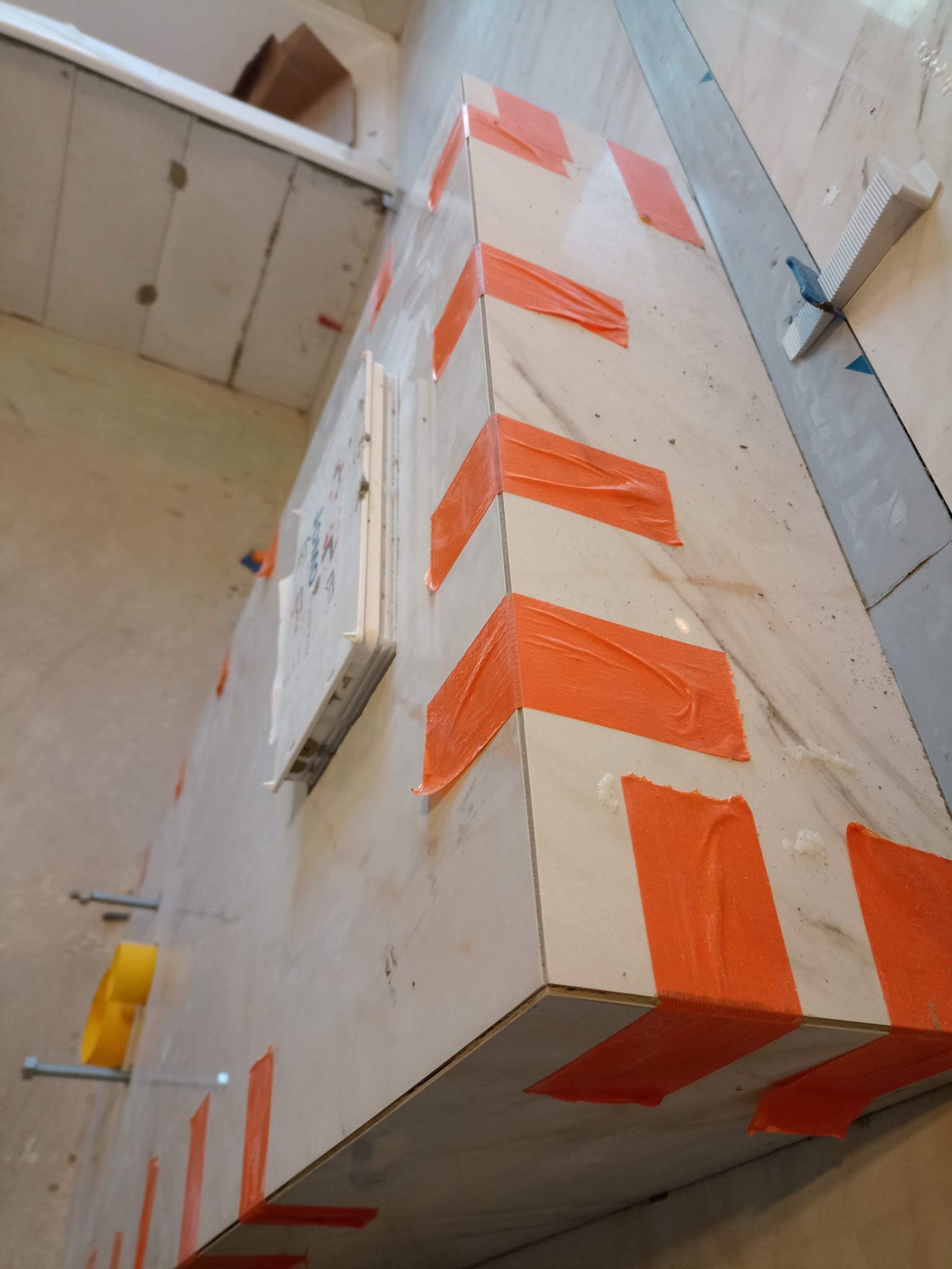 Układanie białych płytek imitujących marmur, zabezpieczonych pomarańczową taśmą malarską na narożniku zabudowy w łazience, widoczne elementy mocujące i poziomica.