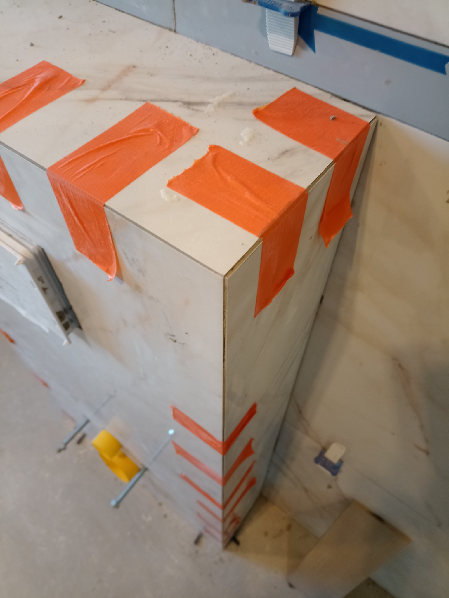Fragment narożnika obłożonego płytkami imitującymi marmur, zabezpieczonego pomarańczową taśmą malarską podczas prac wykończeniowych. Widoczne kliny poziomujące i krzyżyki dystansowe.