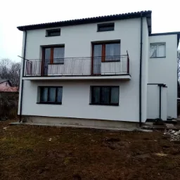 Dwukondygnacyjny dom z otynkowaną, białą elewacją, balkonem z czarną, metalową balustradą i brązowymi ramami okiennymi, otoczony ziemią bez trawy.