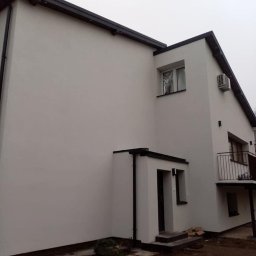 Usługi Ogólnobudowlane Artur Ostrowski - Dwupiętrowy dom z białymi ścianami i czarnym dachem, widok z boku na elewację po renowacji, z klimatyzatorem na ścianie i niezagospodarowanym terenem wokół.