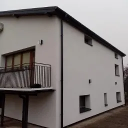 Dwukondygnacyjny dom z balkonem, świeżo otynkowany na biało, z czarną stolarką okienną i rynnami, w pochmurny dzień.