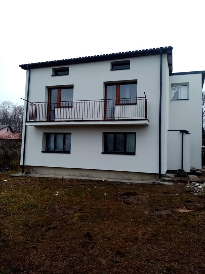 Dwukondygnacyjny dom z otynkowaną, białą elewacją, balkonem z czarną, metalową balustradą i brązowymi ramami okiennymi, otoczony ziemią bez trawy.