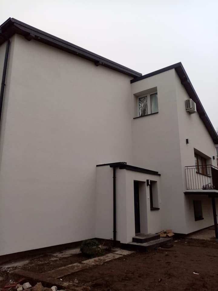 Dwupiętrowy dom z białymi ścianami i czarnym dachem, widok z boku na elewację po renowacji, z klimatyzatorem na ścianie i niezagospodarowanym terenem wokół.