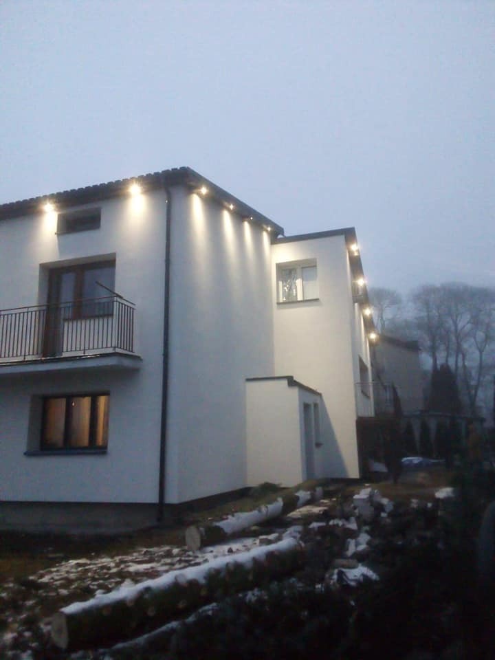 Dwupiętrowy budynek z białymi ścianami i oświetleniem zewnętrznym wzdłuż dachu, widoczny balkon z czarną balustradą i ścięte pnie drzew na pierwszym planie.