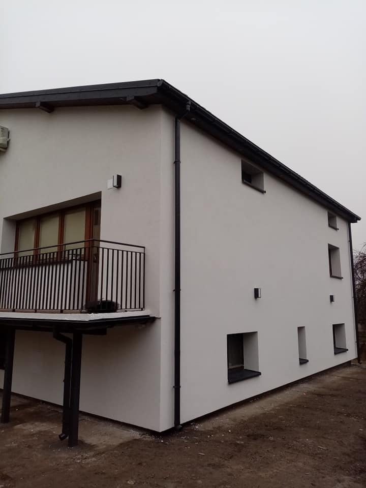 Dwukondygnacyjny dom z balkonem, świeżo otynkowany na biało, z czarną stolarką okienną i rynnami, w pochmurny dzień.