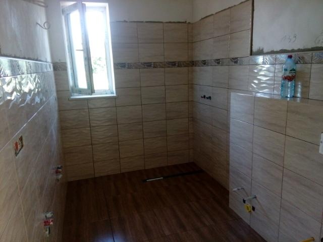 Wnętrze łazienki w trakcie remontu, z ułożonymi beżowymi płytkami na ścianach, imitującymi fale, brązowymi panelami na podłodze i fragmentem nieotynkowanej ściany nad płytkami, butelka wody...