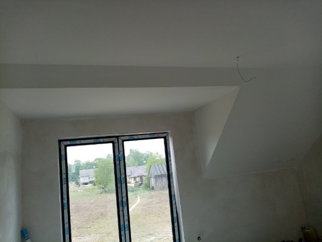 Wnętrze pokoju na poddaszu w trakcie wykańczania, widoczne surowe ściany po szpachlowaniu, okno balkonowe z widokiem na wiejski krajobraz, wystający kabel z sufitu.
