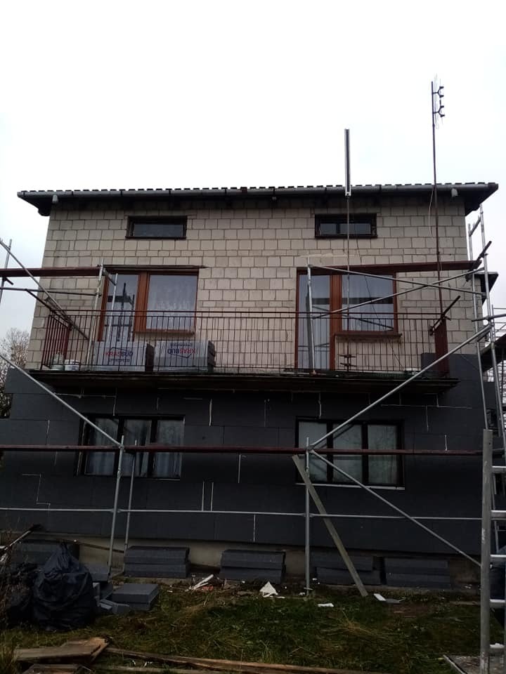 Surowy, dwukondygnacyjny budynek z białej cegły w trakcie ocieplania elewacji, widoczne rusztowania, czarne płyty izolacyjne na dolnej kondygnacji, okna zabezpieczone folią.