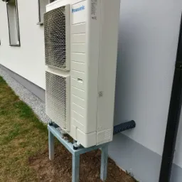 Zewnętrzna jednostka pompy ciepła Panasonic zamontowana na metalowym stelażu przy ścianie budynku z widocznym odprowadzeniem skroplin.