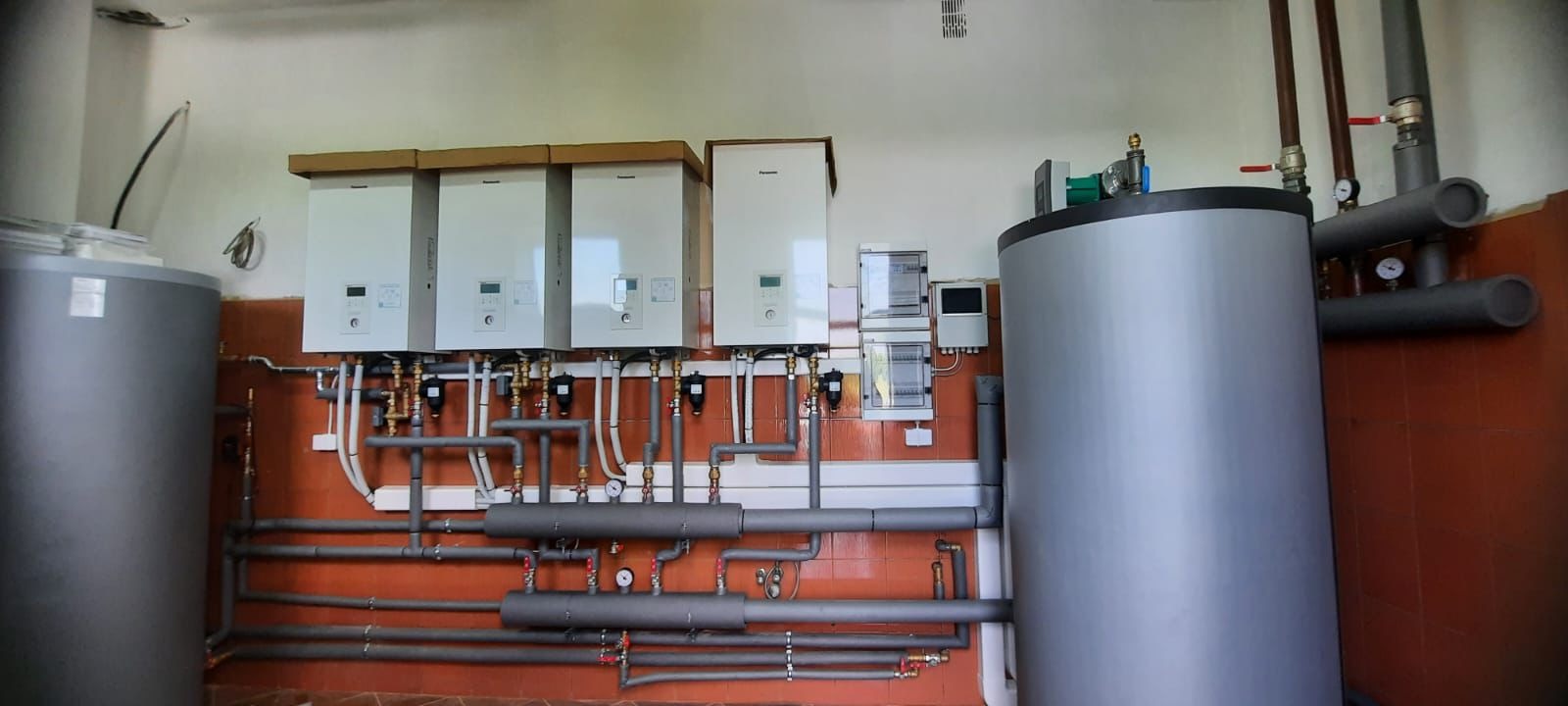 Kaskada czterech pomp ciepła Panasonic 16kW T-CAP w kotłowni, z widocznymi zbiornikami buforowymi, orurowaniem i osprzętem elektrycznym na pomarańczowo-białej ścianie.