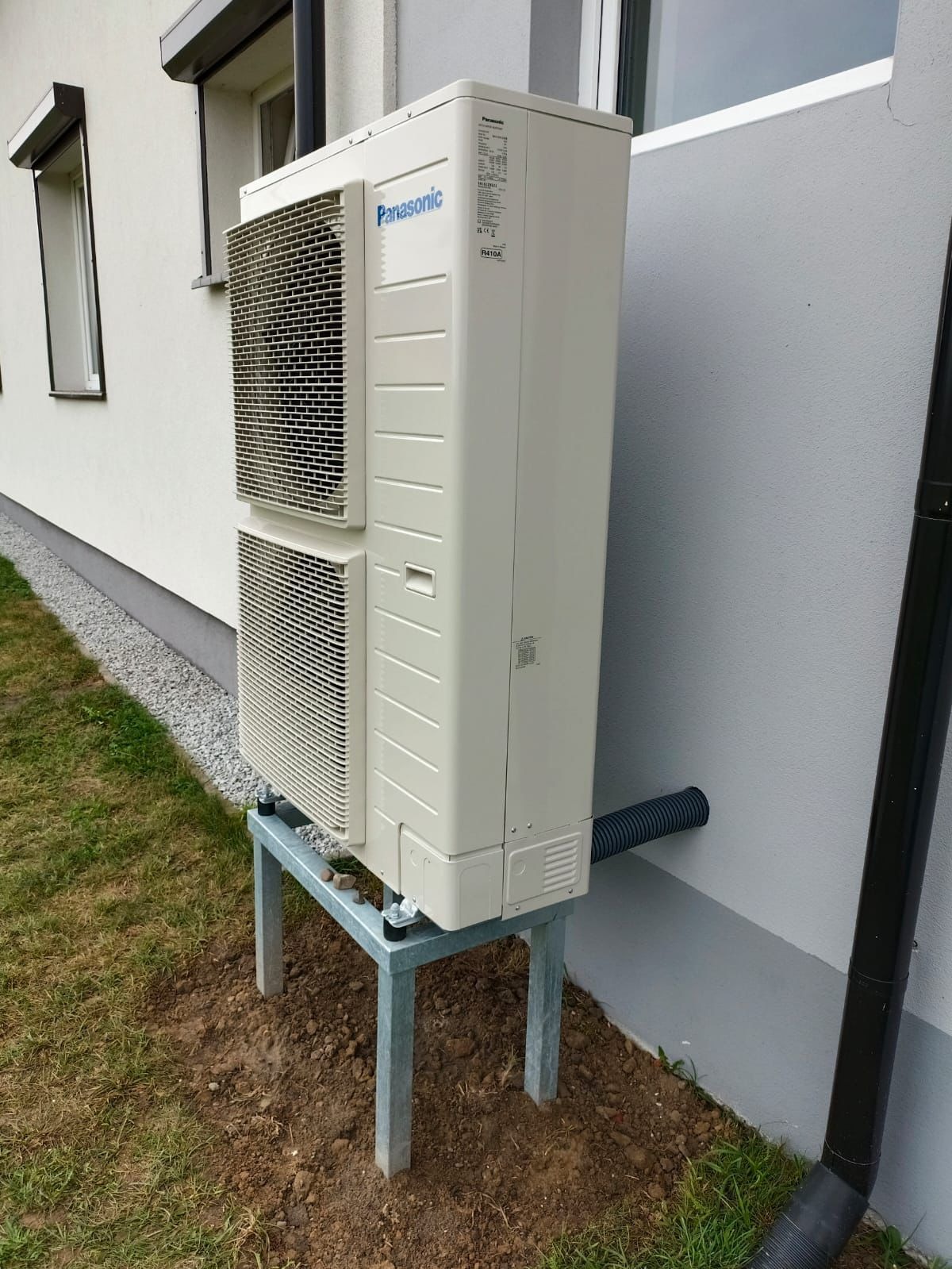 Zewnętrzna jednostka pompy ciepła Panasonic zamontowana na metalowym stelażu przy ścianie budynku z widocznym odprowadzeniem skroplin.