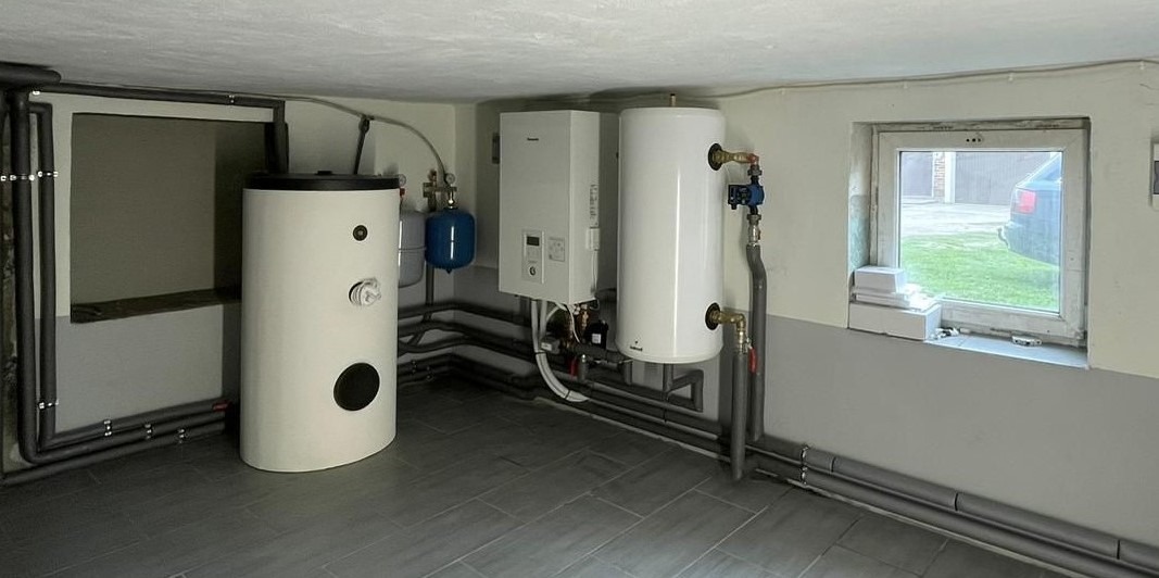 Widok kotłowni z dwoma białymi zbiornikami, jednostką sterującą na ścianie, niebieskim zbiornikiem wyrównawczym oraz szarymi rurami biegnącymi wzdłuż ścian i podłogi z szarych płytek. W tle...