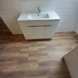 Nowoczesna łazienka z białą umywalką nablatową na szafce z trzema szufladami, białe płytki na ścianach ułożone w cegiełkę, podłoga wyłożona panelami imitującymi drewno, lustro prostokątne...