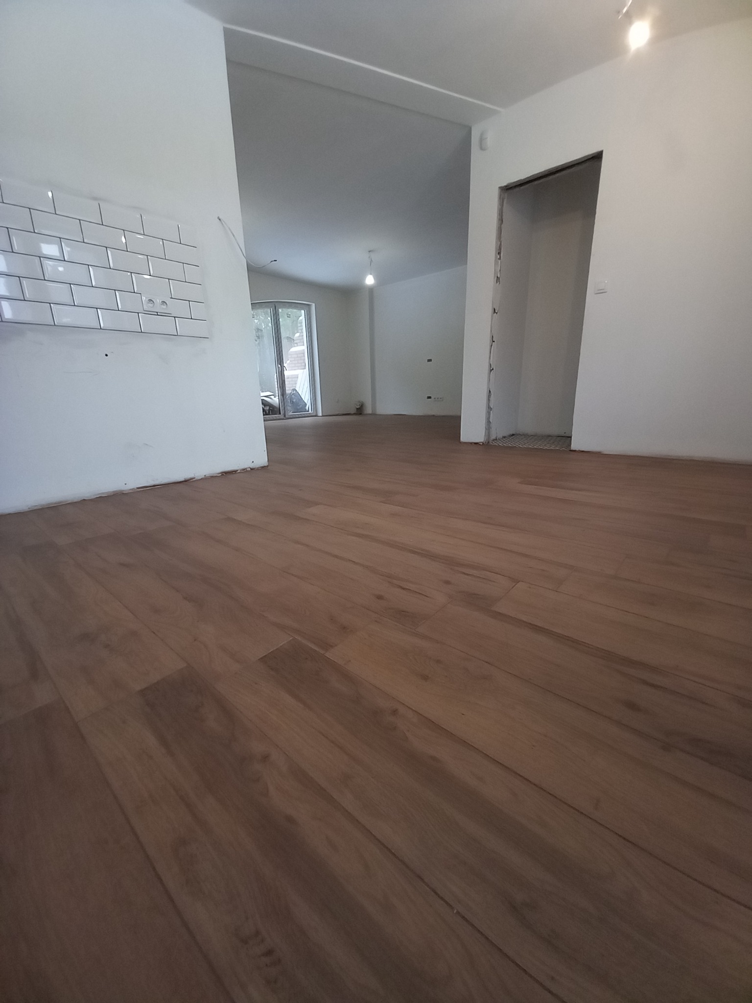 Ukończona podłoga z paneli drewnopodobnych w jasnym, odnowionym wnętrzu. Widoczne białe ściany z fragmentem glazury w stylu 'metro' oraz otwór drzwiowy bez ościeżnicy.