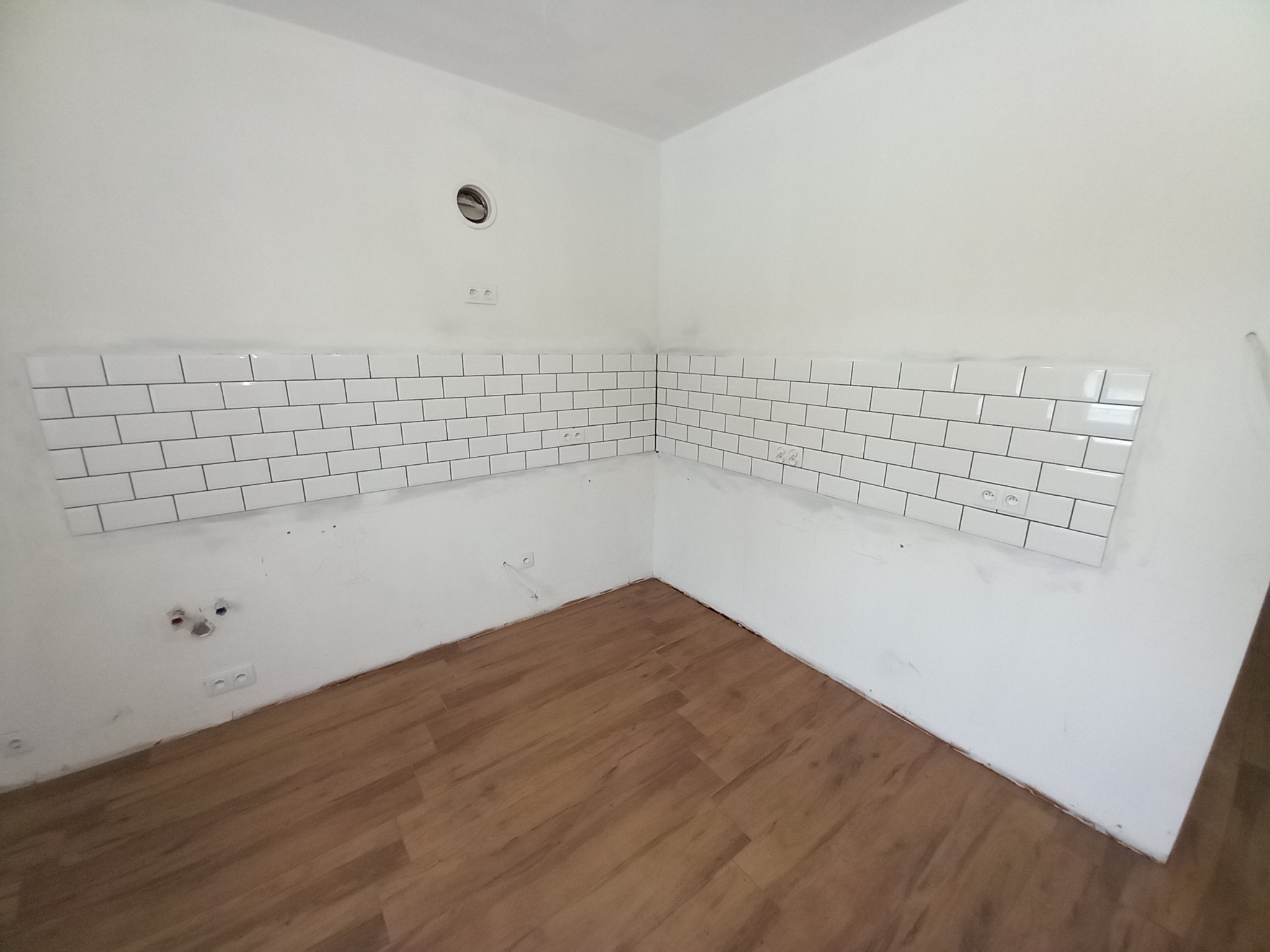 Fragment kuchni w trakcie układania białych, prostokątnych płytek na ścianie, widoczne niedoskonałości i ślady po pracach, drewnopodobna podłoga.