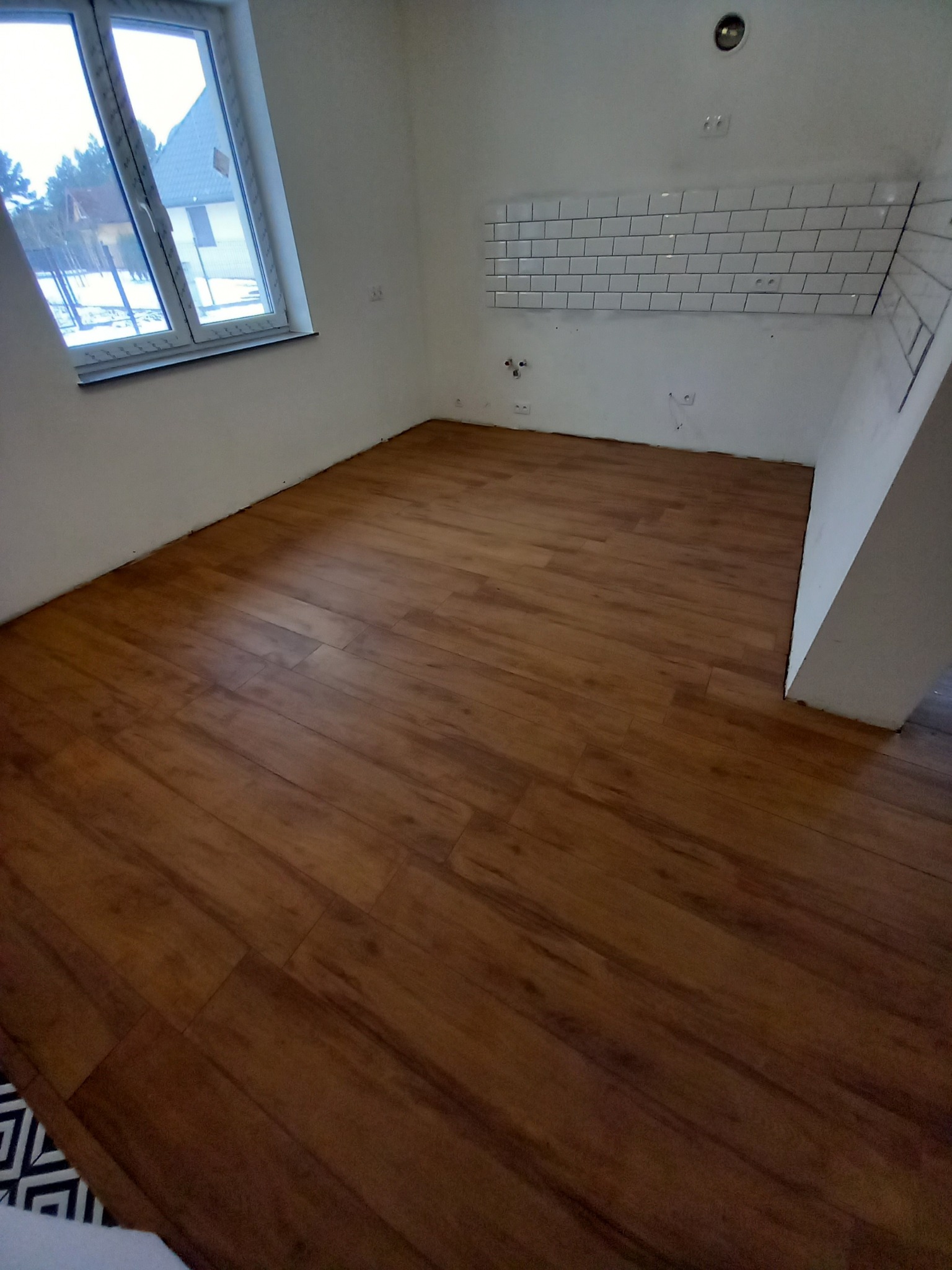 Wnętrze po remoncie z drewnopodobną podłogą, fragmentem ułożonych białych płytek nad blatem, oknem z widokiem na dom i zimowy krajobraz.