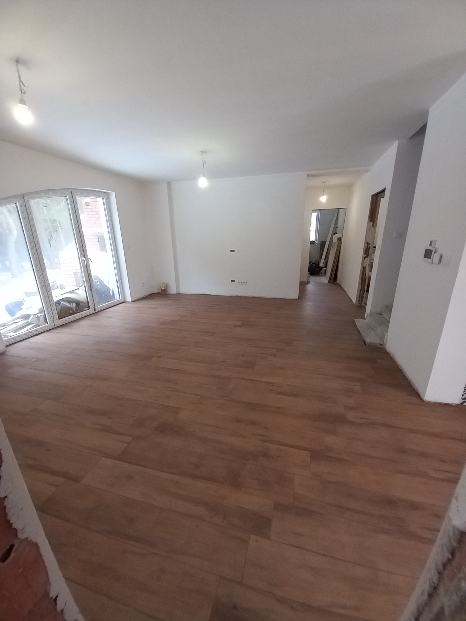 Pusty pokój po remoncie z nową podłogą z paneli imitujących drewno, świeżo pomalowane ściany na biało, widoczne gniazdka elektryczne i okno balkonowe z widokiem na ceglany budynek.