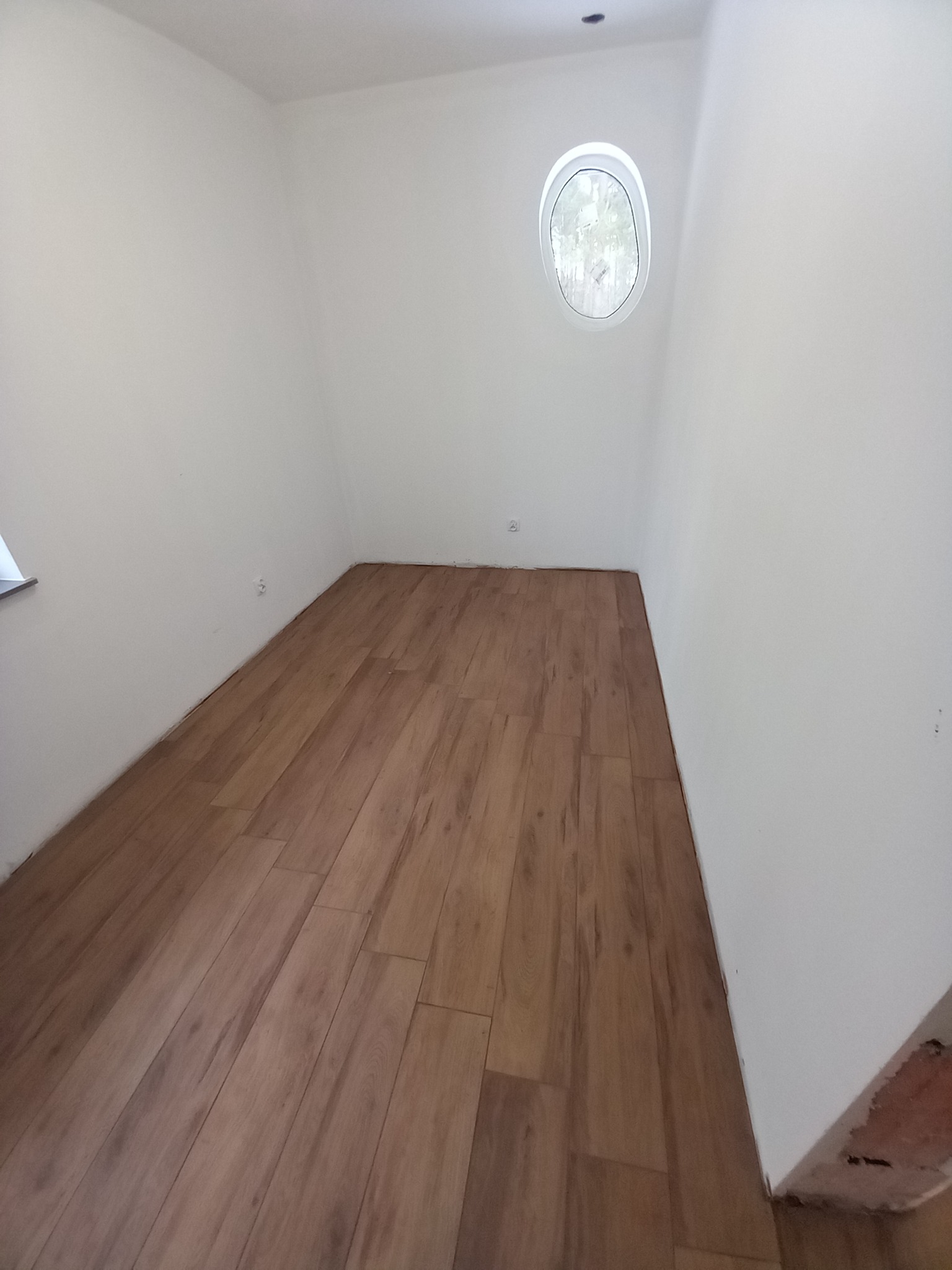 Poddasze po remoncie z drewnopodobną podłogą, okrągłym oknem i świeżo pomalowanymi na biało ścianami, widoczne gniazdka elektryczne.