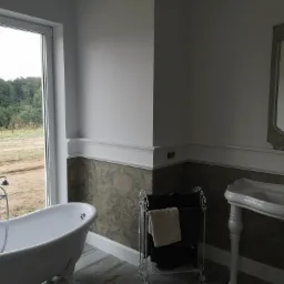 Elegancka łazienka z wolnostojącą wanną przy oknie z widokiem na zieleń, umywalką na ozdobnych nogach i dekoracyjną tapetą w dolnej części ściany.