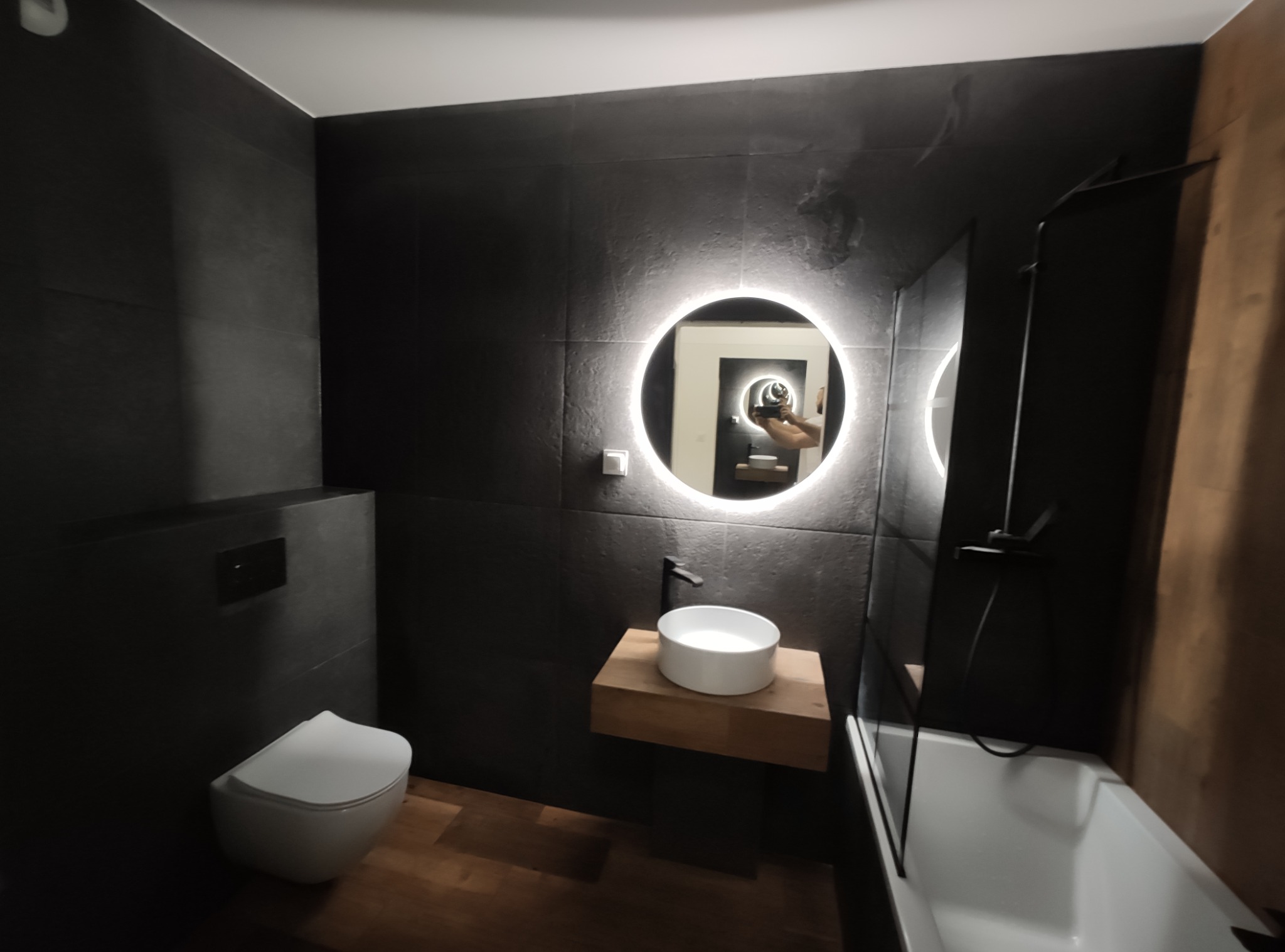 Nowoczesna łazienka z czarnymi płytkami, okrągłym lustrem z podświetleniem LED, wiszącą białą umywalką na drewnianym blacie i białą wanną z czarną armaturą.