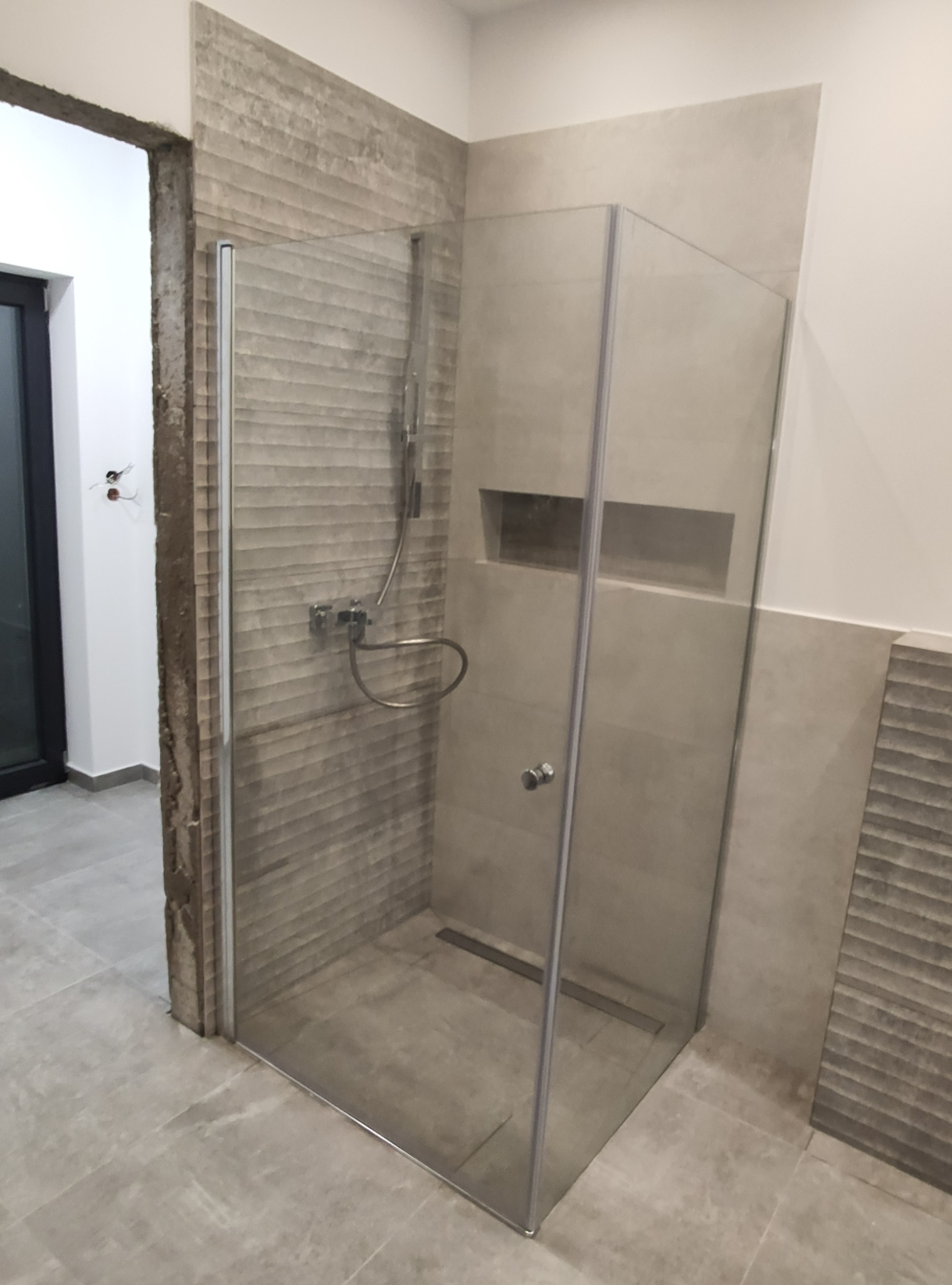 Nowoczesna kabina prysznicowa z szarymi płytkami o fakturze drewna, z odpływem liniowym i wnęką w ścianie, widoczna na tle surowego otworu drzwiowego.