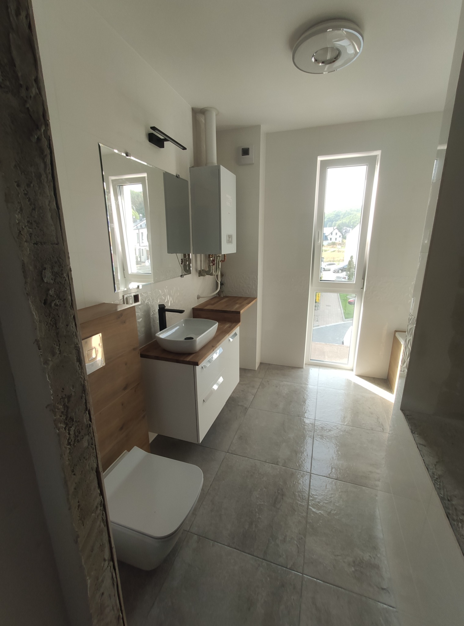 Nowoczesna łazienka z wiszącą toaletą, umywalką nablatową na szafce z drewnianym blatem, lustrem z oświetleniem LED i grzejnikiem gazowym, wykończona szarą płytką podłogową i białymi ścianami...