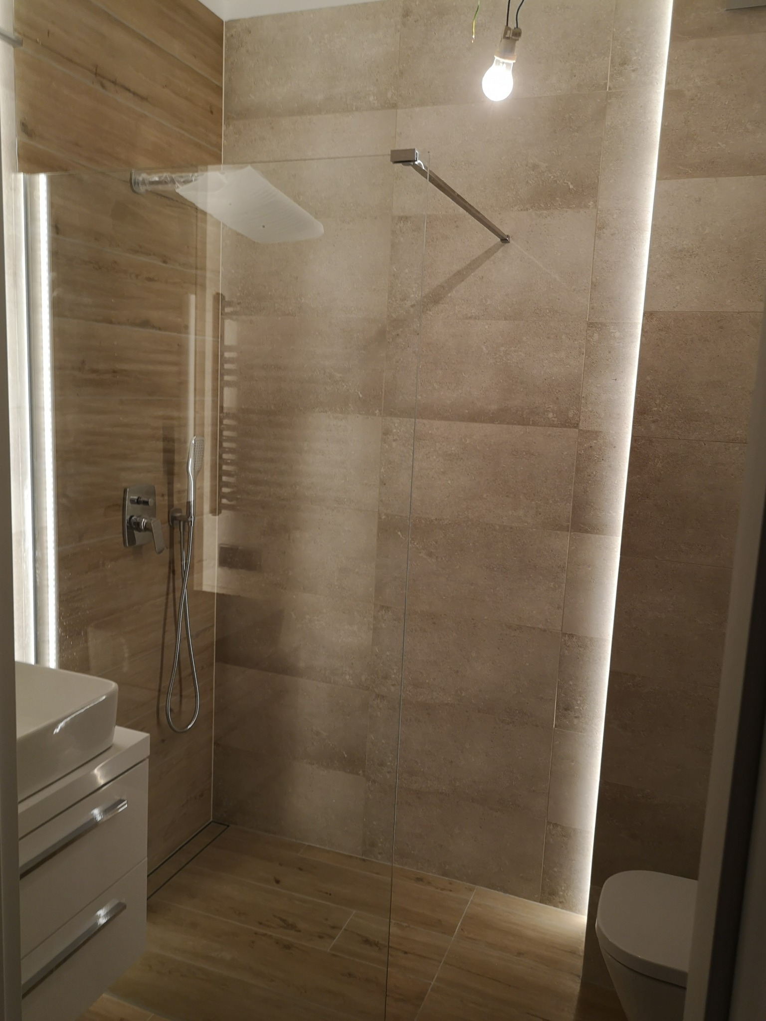 Nowoczesna kabina prysznicowa z szklaną ścianką, baterią podtynkową i odpływem liniowym, wykończona płytkami w odcieniach beżu i drewna, z oświetleniem LED wzdłuż ściany i wiszącą żarówką.