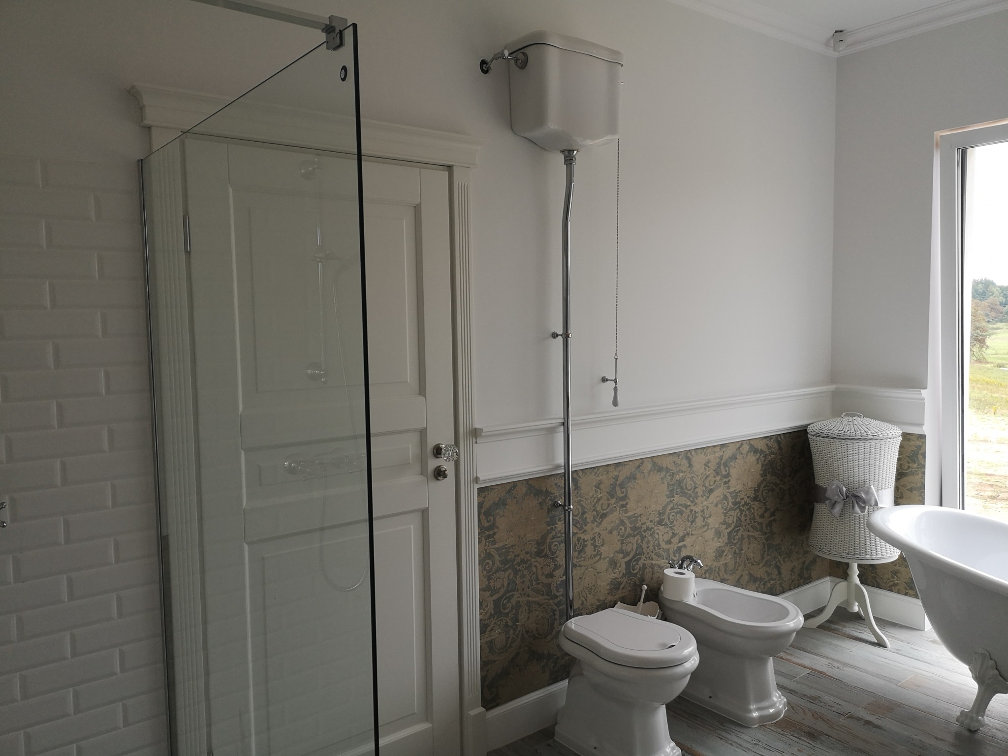 Klasyczna łazienka z białą armaturą: toaleta z wysokim zbiornikiem, bidet, wanna na nóżkach i przeszklona kabina prysznicowa. Ściany zdobią białe płytki w stylu metro i tapeta w kwiatowe wzory.