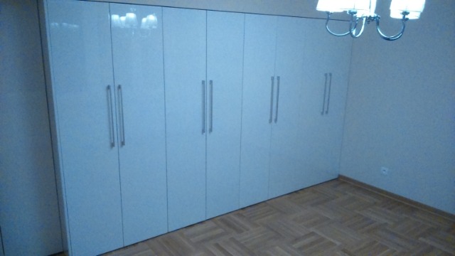 Biała, błyszcząca szafa wnękowa z długimi, pionowymi uchwytami, zajmująca całą ścianę w pokoju z parkietem ułożonym w jodełkę.