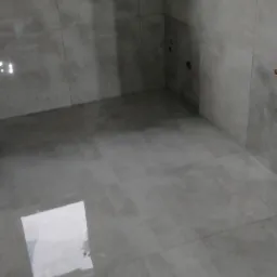 Świeżo położone szare płytki podłogowe w łazience, z widocznym odbiciem okna w połysku płytek i przygotowanymi otworami w ścianie na armaturę.