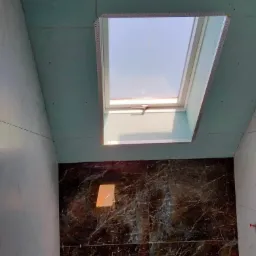 Wnętrze łazienki w trakcie montażu sufitu z płyty kartonowo-gipsowej, widoczne okno dachowe oraz częściowo wykończone ściany z marmurowymi płytkami w odcieniach brązu i bieli.