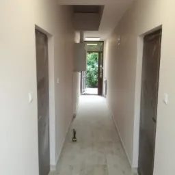 Korytarz w trakcie remontu z położonymi płytkami podłogowymi, widoczne drzwi w surowym stanie, w tle otwór drzwiowy z widokiem na zieleń, na podłodze wiertarka akumulatorowa.