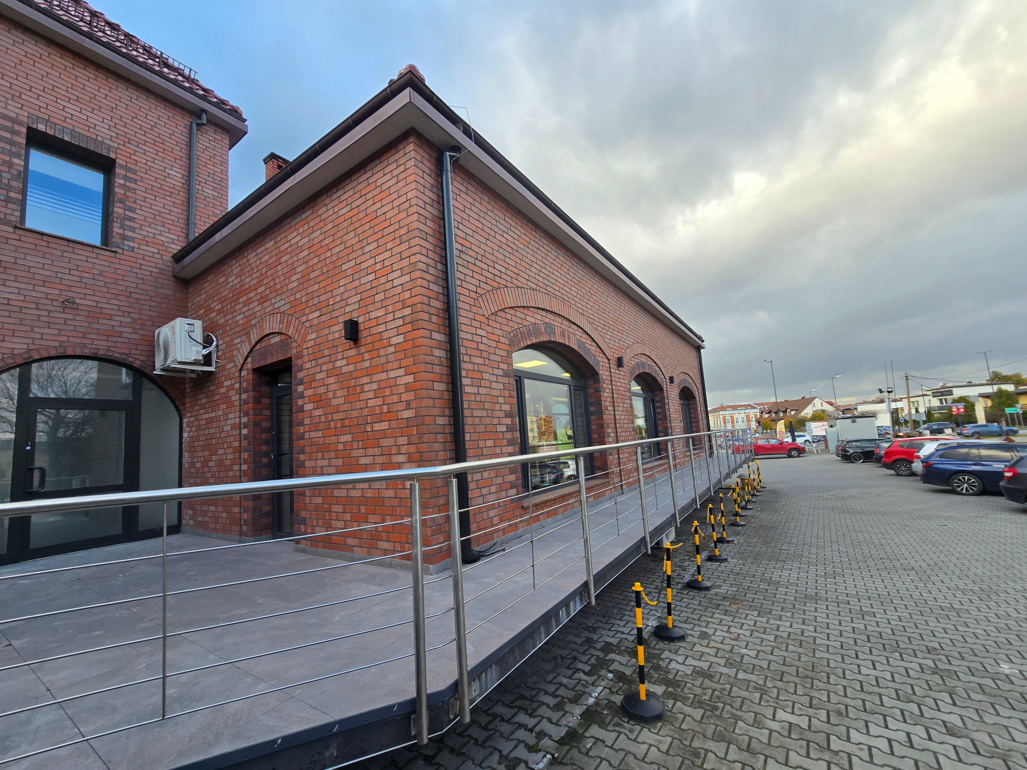 Budynek z czerwonej cegły z rampą dla wózków inwalidzkich i parkingiem. Widoczne detale architektoniczne, takie jak łukowe okna i metalowe balustrady.