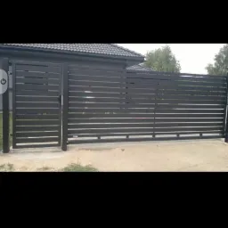 Nowoczesna, pozioma brama wjazdowa i przęsła ogrodzeniowe z metalowych profili, pomalowane na ciemny kolor, z widocznym fragmentem domu w tle.