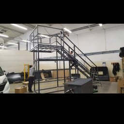 P.P.H.U Exomet - Konstrukcja metalowa z platformą i schodami w hali produkcyjnej, dwóch pracowników przy pracy, oświetlenie sufitowe.