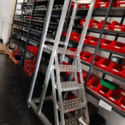 Aluminiowa drabina magazynowa z czterema stopniami o antypoślizgowej powierzchni, na kółkach, ustawiona przed regałami z czerwonymi i czarnymi pojemnikami na drobne elementy.