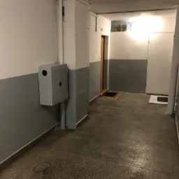 Korytarz biurowy z szarą lamperią, widoczną skrzynką elektryczną na ścianie, betonową podłogą i drzwiami w tle.