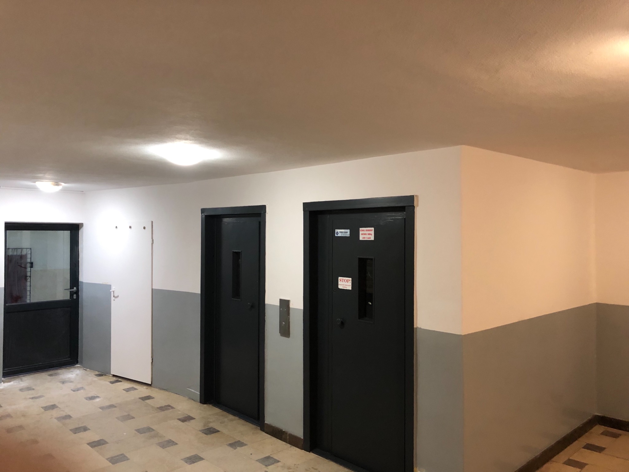 Korytarz w Stąporkowie z dwoma czarnymi drzwiami do windy, pomalowany na biało i szaro, z widocznym sufitem i podłogą w kratkę.