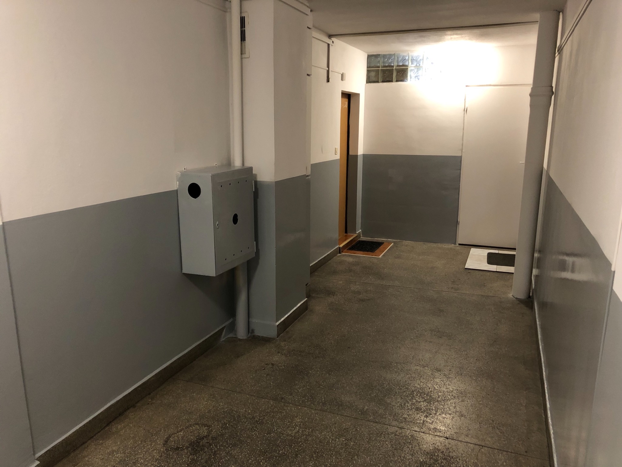Korytarz biurowy z szarą lamperią, widoczną skrzynką elektryczną na ścianie, betonową podłogą i drzwiami w tle.
