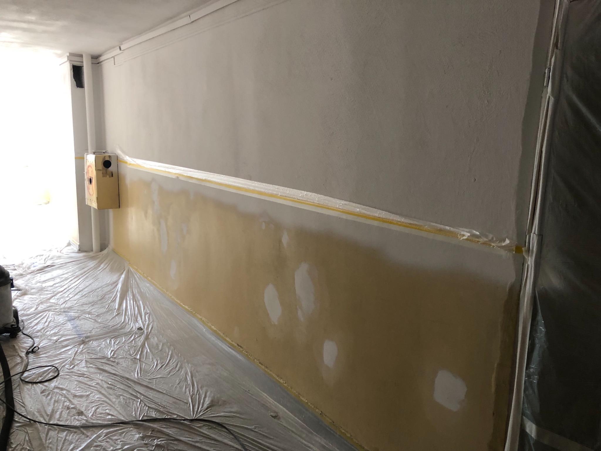 Przygotowanie ściany do malowania: ściana z żółtym pasem zabezpieczającym, zagruntowane białe plamy, folia ochronna na podłodze, widoczny odkurzacz przemysłowy.