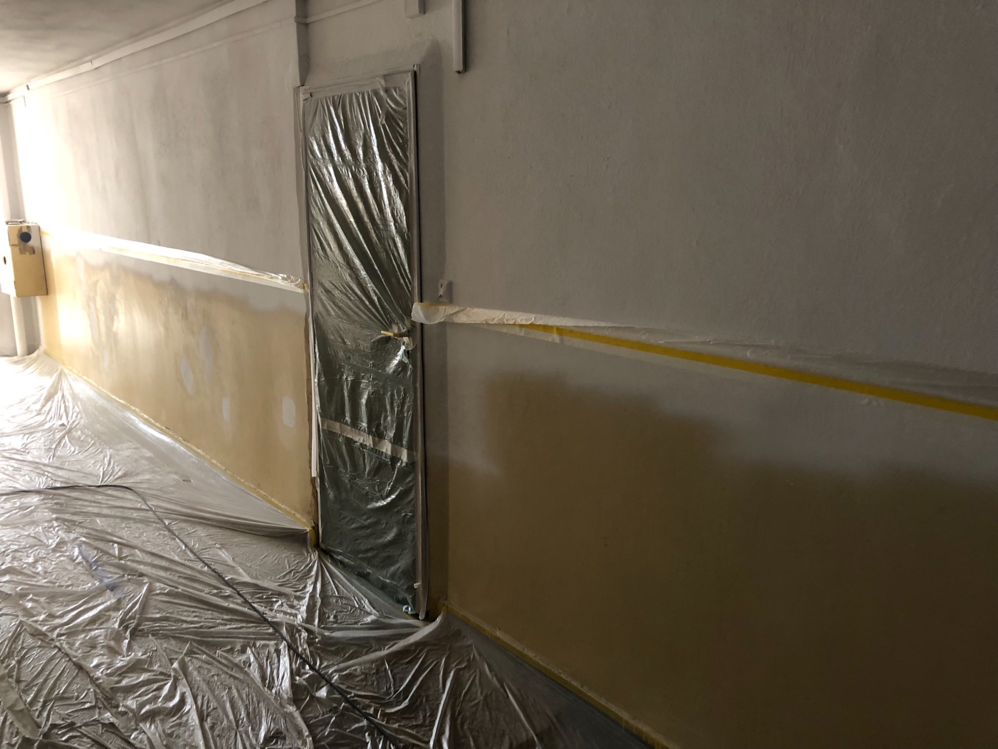 Przygotowanie do malowania biura: ściany zabezpieczone folią i taśmą malarską, drzwi osłonięte folią ochronną, widoczne ślady po szpachlowaniu na ścianie.