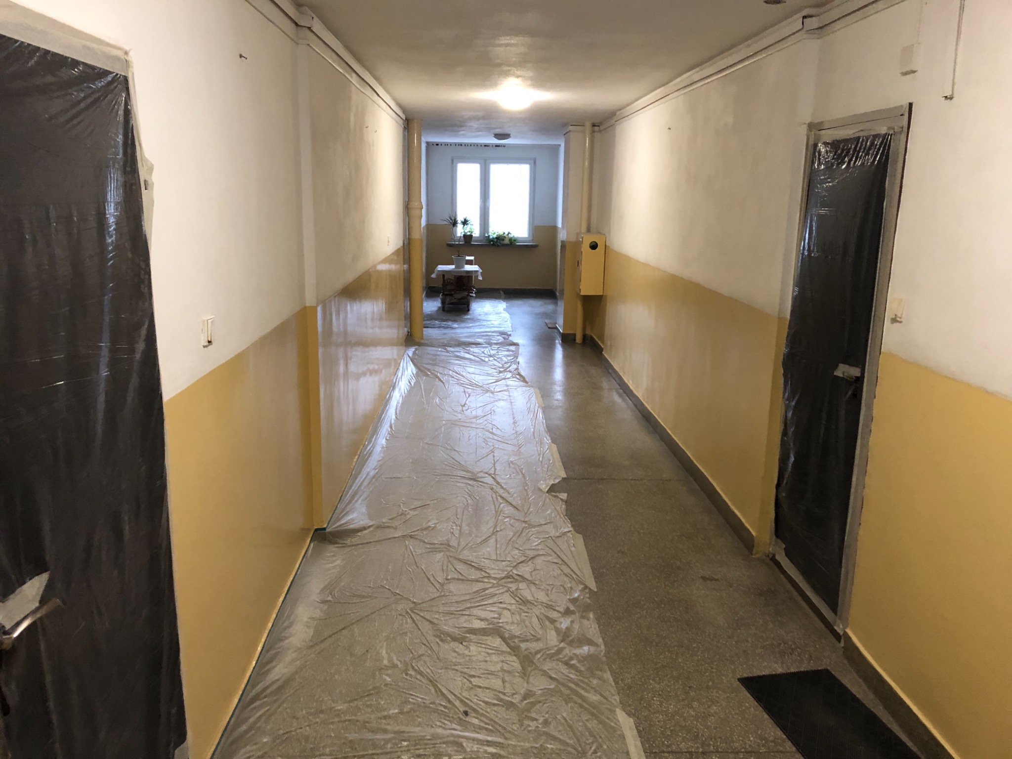 Korytarz w bloku zabezpieczony folią malarską przed malowaniem ścian na biało i żółto, drzwi oklejone folią ochronną, widoczne ślady taśmy malarskiej.