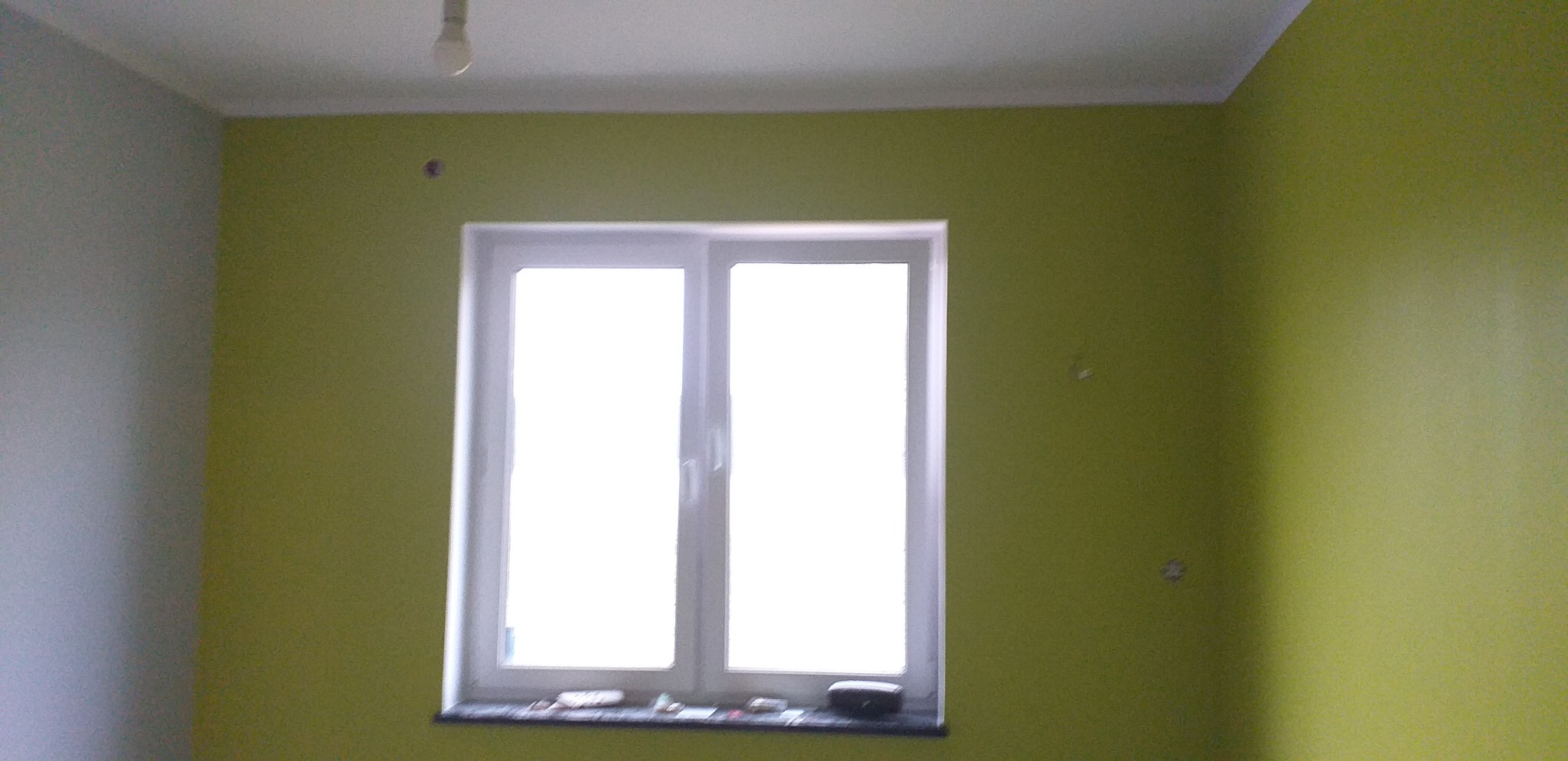 Pokój po świeżym malowaniu na kolor limonkowy, z widocznymi drobnymi niedoskonałościami na ścianie i oknem z białymi ramami, na parapecie leżą drobne przedmioty.