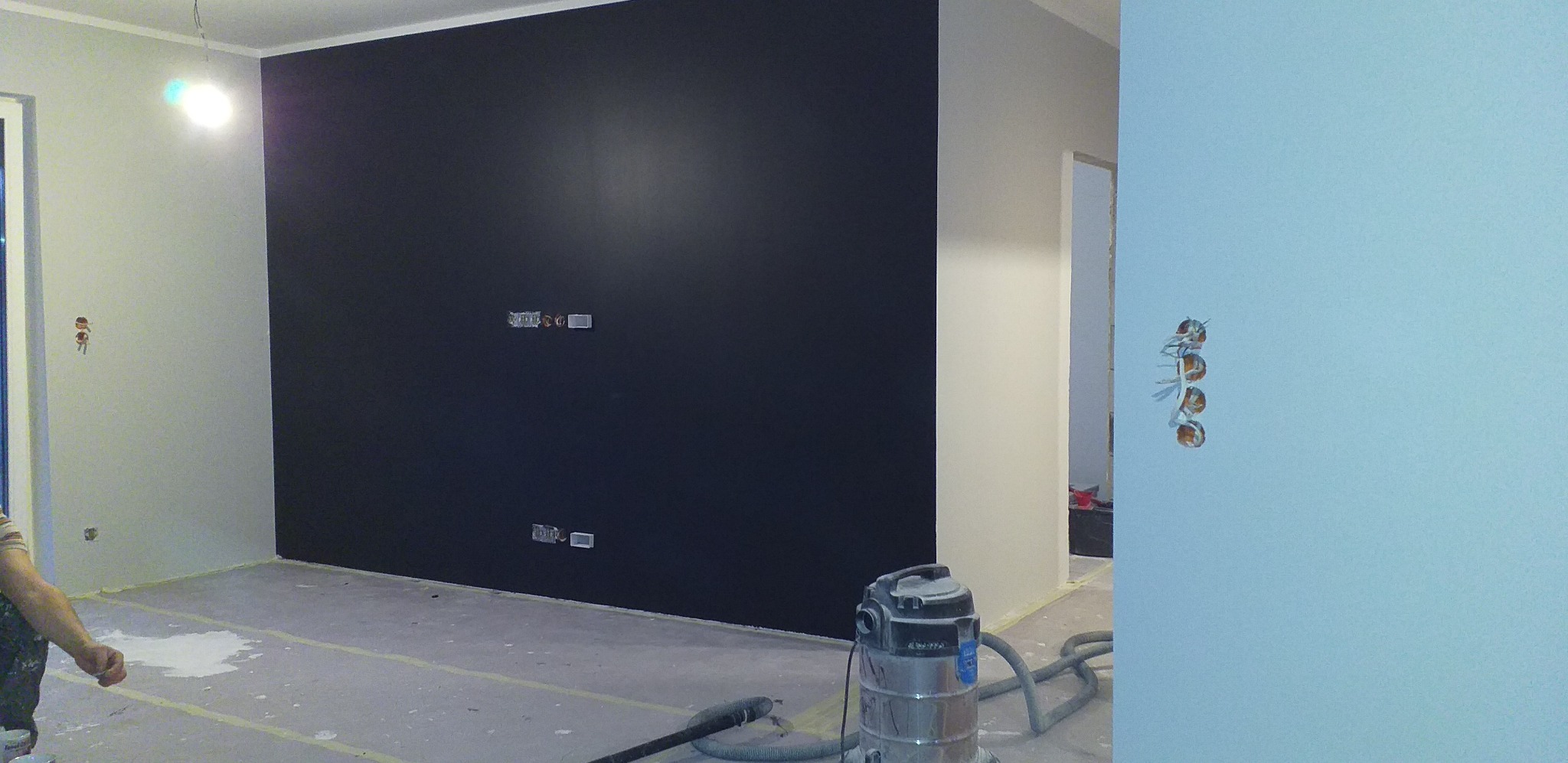 Pokój w trakcie malowania: ściana pomalowana na czarno, widoczne puszki elektryczne, zabezpieczona podłoga, odkurzacz przemysłowy. Fragment malarza w lewym dolnym rogu.