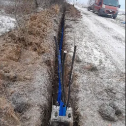 Wykop z ułożonymi kablami elektrycznymi w niebieskich rurach ochronnych, widoczna skrzynka betonowa z mocowaniami, w tle pojazd dostawczy na ośnieżonej drodze.