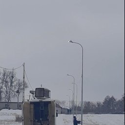 MOTOKOP - Pracownik w zimowym krajobrazie, montujący oświetlenie uliczne, z otwartym busem serwisowym w tle.