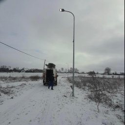 MOTOKOP - Montaż oświetlenia ulicznego w zimowej scenerii: pracownik w oddali przy furgonetce, świeżo postawiona latarnia z włączonym światłem na zasypanym śniegiem terenie.