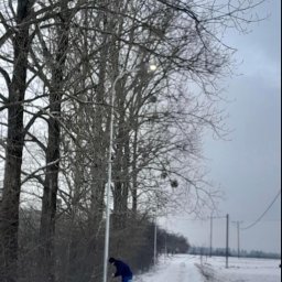 MOTOKOP - Pracownik w niebieskim kombinezonie przy słupie oświetleniowym w zimowym krajobrazie, widoczna ośnieżona droga i drzewa bez liści.
